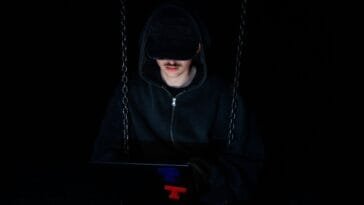 Rahapelihuijaus verkossa – huppupäinen hakkeri käyttämässä tietokonetta pimeässä